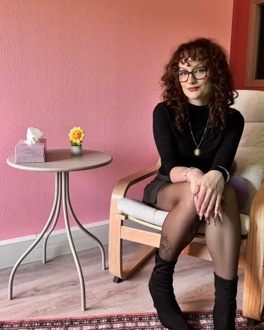 Portrait de la sophrologue dans son cabinet à Metz, assise dans un fauteuil devant un mur rose, à côté d'une petite table avec un tournesol en crochet